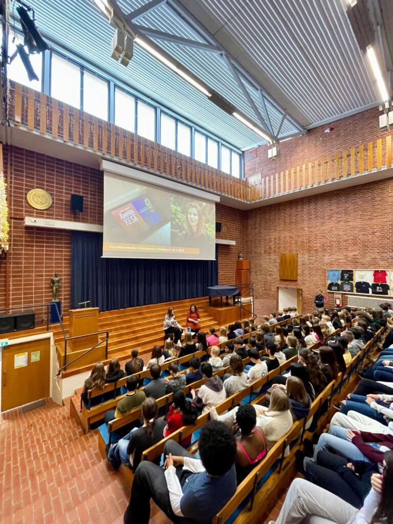 Kuvassa kirjailija Taina Niemi ja Kulosaaren yhteiskoulun kieltenopettaja Sanni Taskinen, Keskushallissa. Yleisössä on oppilaita kuuntelemassa haastattelua.