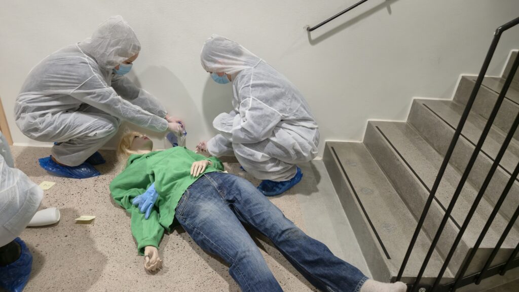 Rikostekninen tutkimus:
Opiskelija tutkimassa rikospaikkaa valkoinen suojapuku yllään. 
