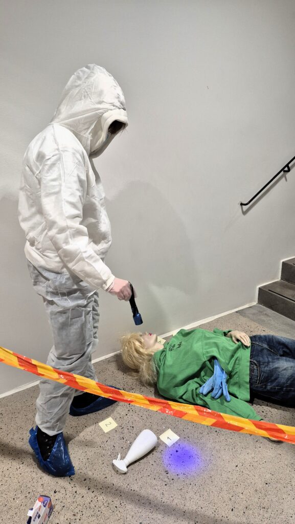 Rikostekninen tutkimus:
Opiskelija tutkimassa rikospaikkaa valkoinen suojapuku yllään.  Osoittaa UV-valolla lattiaan.
